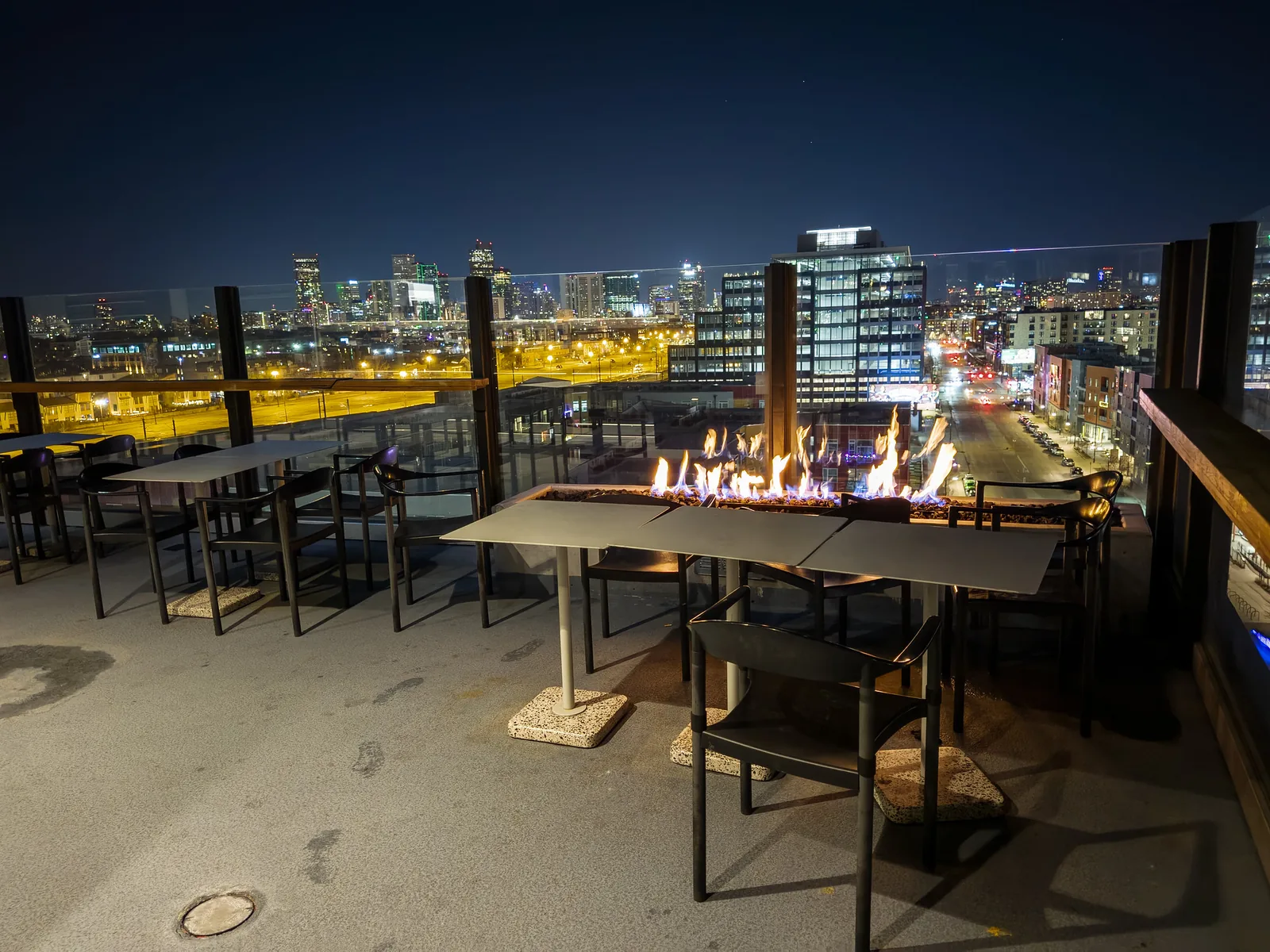 Tables around a lit fire pit on a restaurant's rooftop patio, with a city skyline beyond at night
