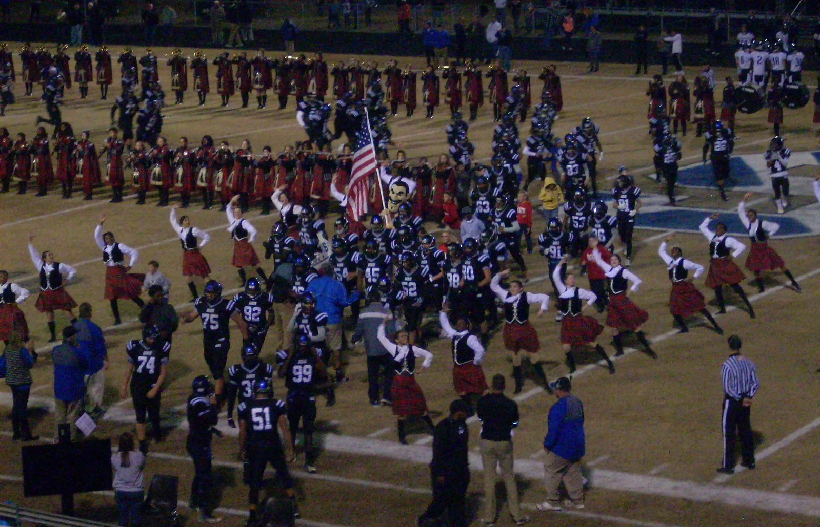 A high school football team runs onto the field through its marching band and dance line at a night game