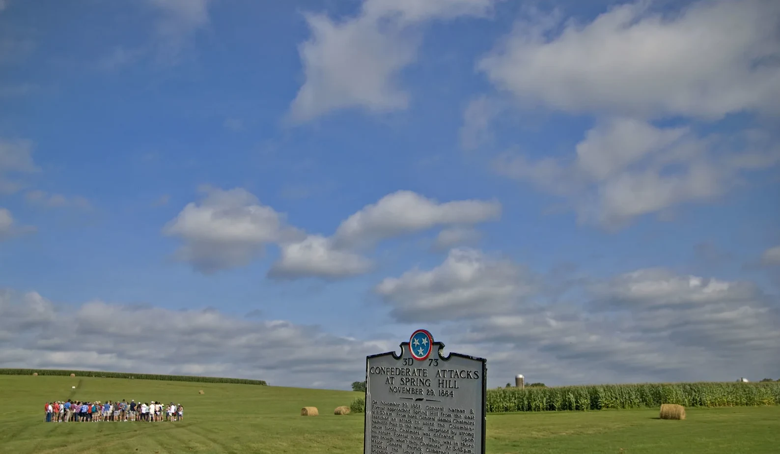 A Tennessee Historical Commission marker for the Confederate attacks at Spring Hill on the preserved battlefield, with a tour group in the field beyond