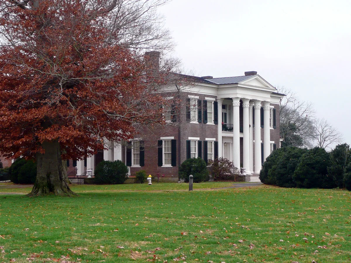 Rippa Villa's brick front and white two-story portico behind a lawn and a red-leaved oak in fall on Main Street in Spring Hill