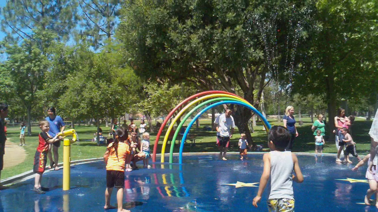 Families and children play under a rainbow arch at a park splash pad on Memorial Day